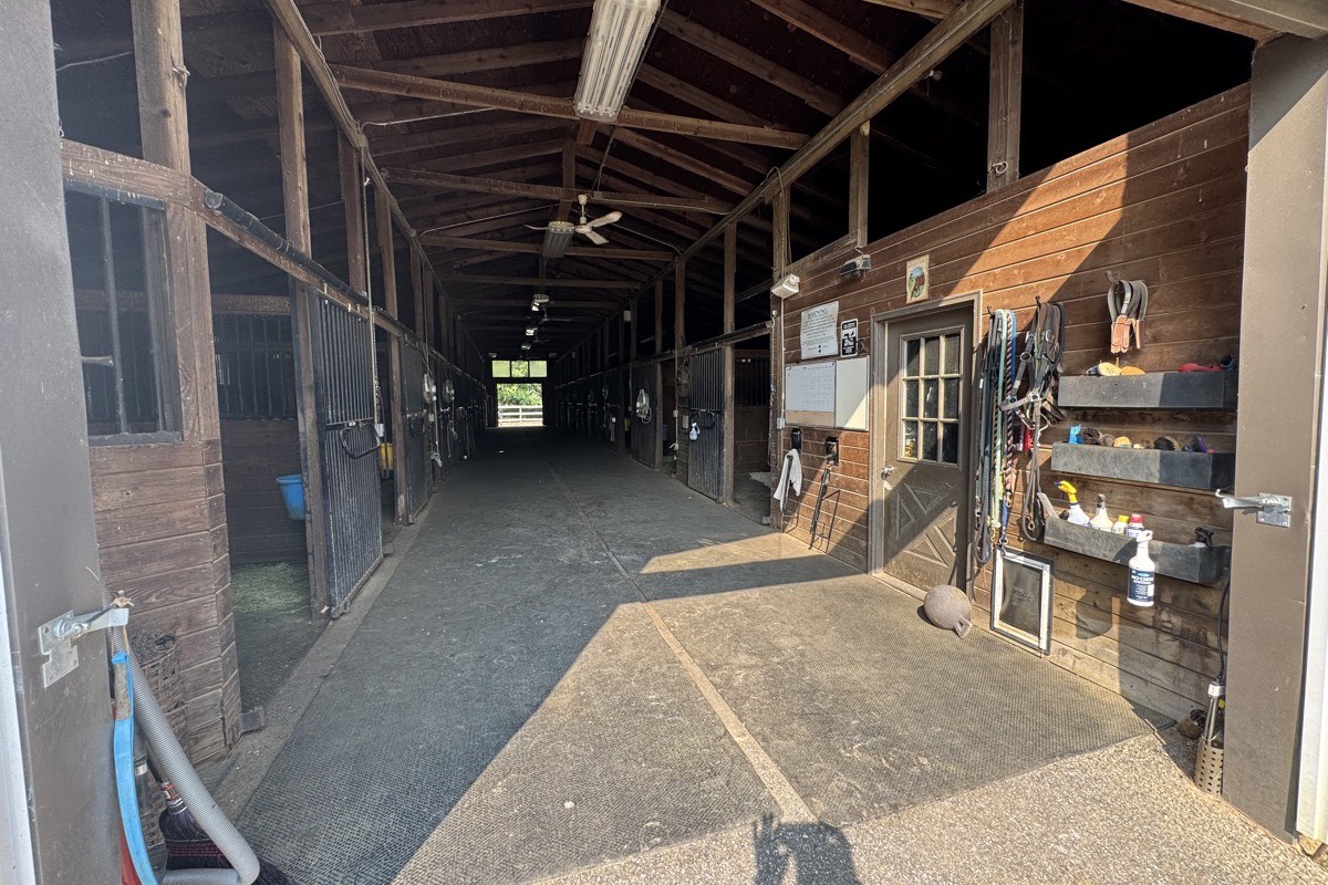 Looking down the main barn aisle, stall fronts on both sides and tack hanging by the office door